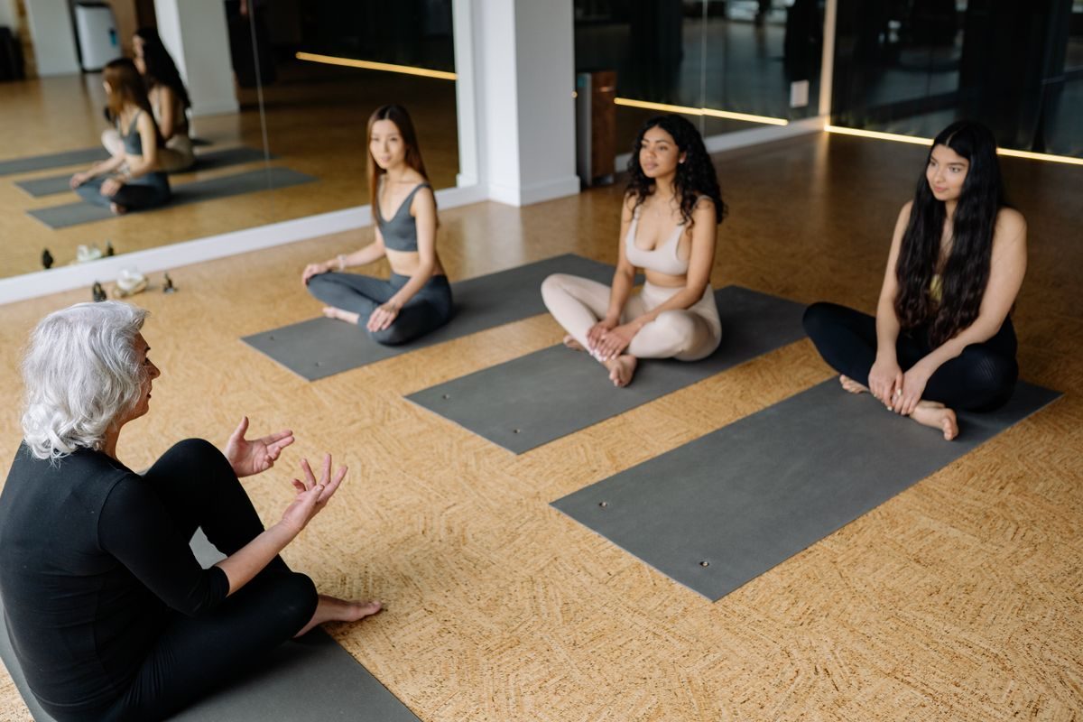 A senior woman sitting on a yoga mat talking with younger students in a studio